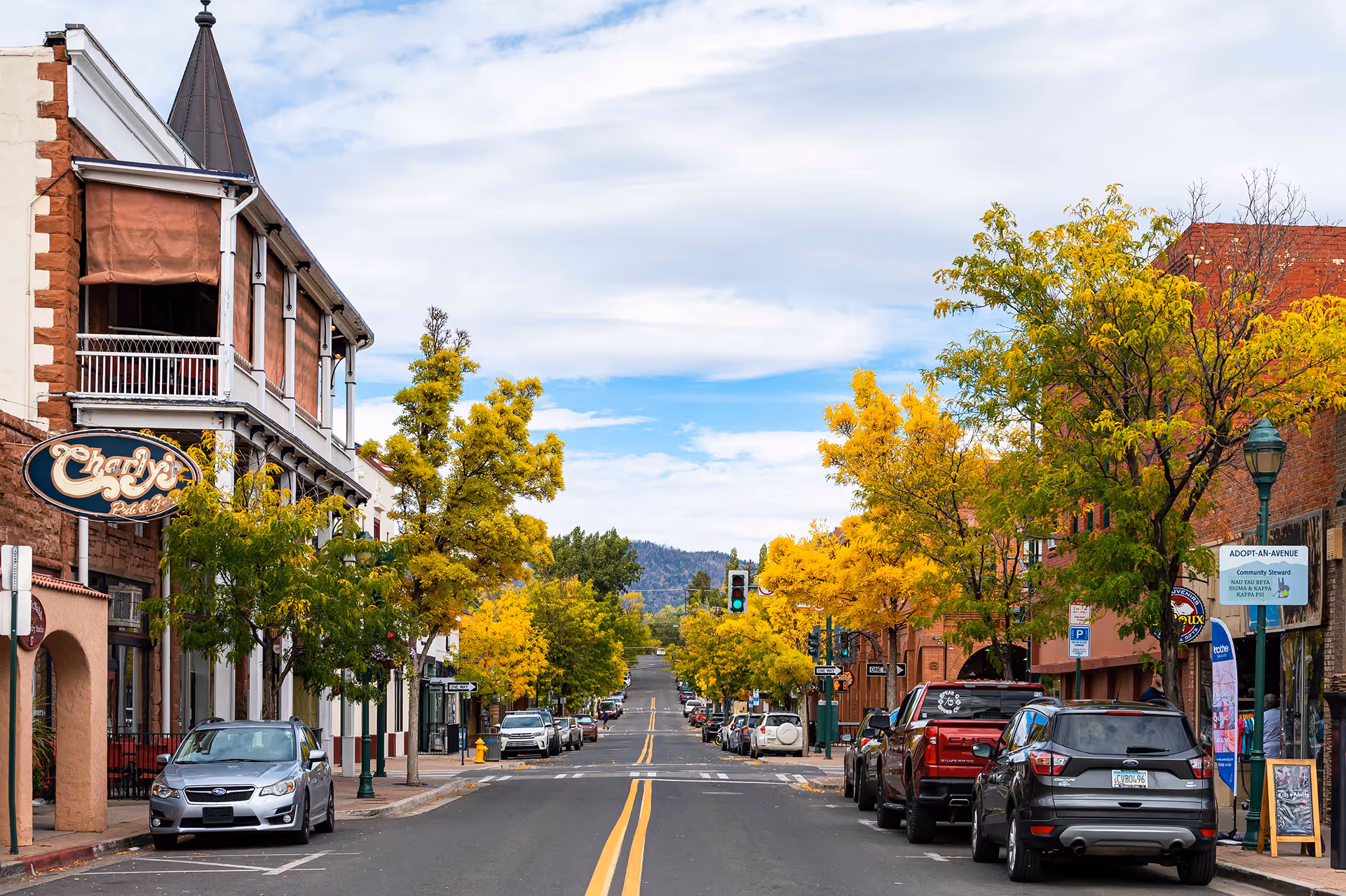 Street view of a small town lined with yellow autumn trees, parked cars, and historic brick buildings under a partly cloudy sky.